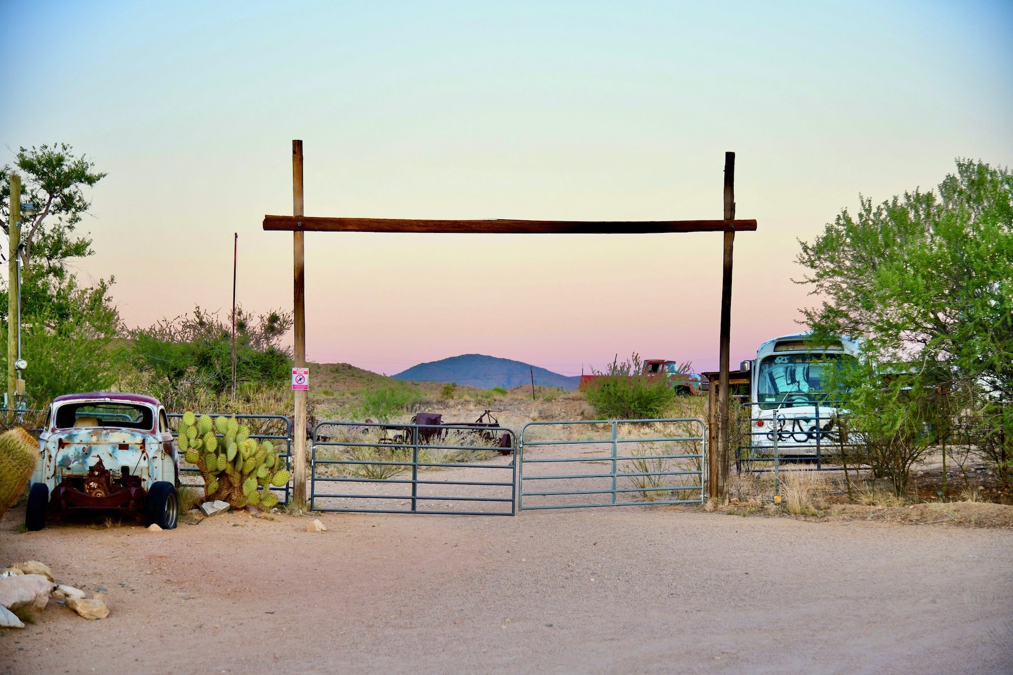 Gate in desert at dusk between abandoned car and bus